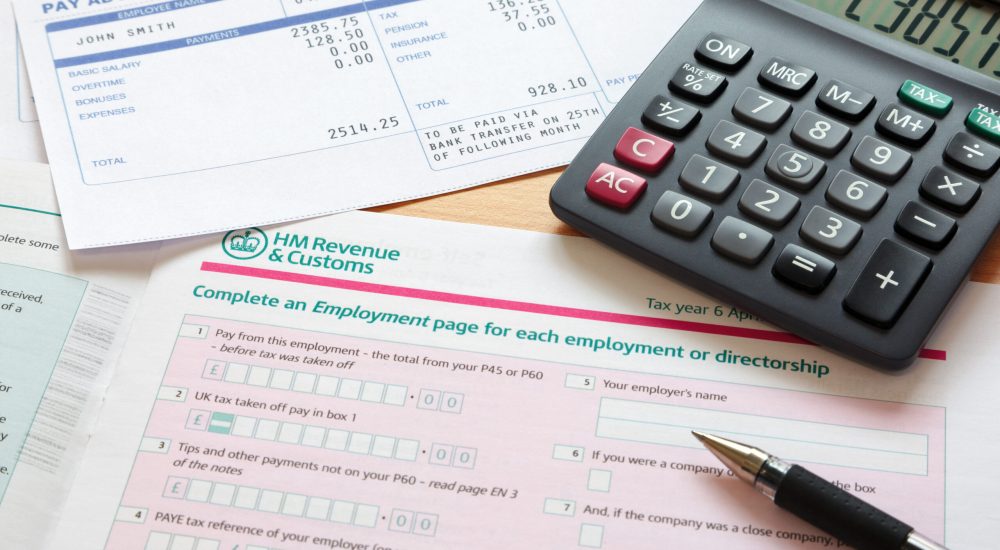 Photo of a UK self assessment tax return with calculator and payslips. The payslip is a mock up the names and all other information on it is fictional.