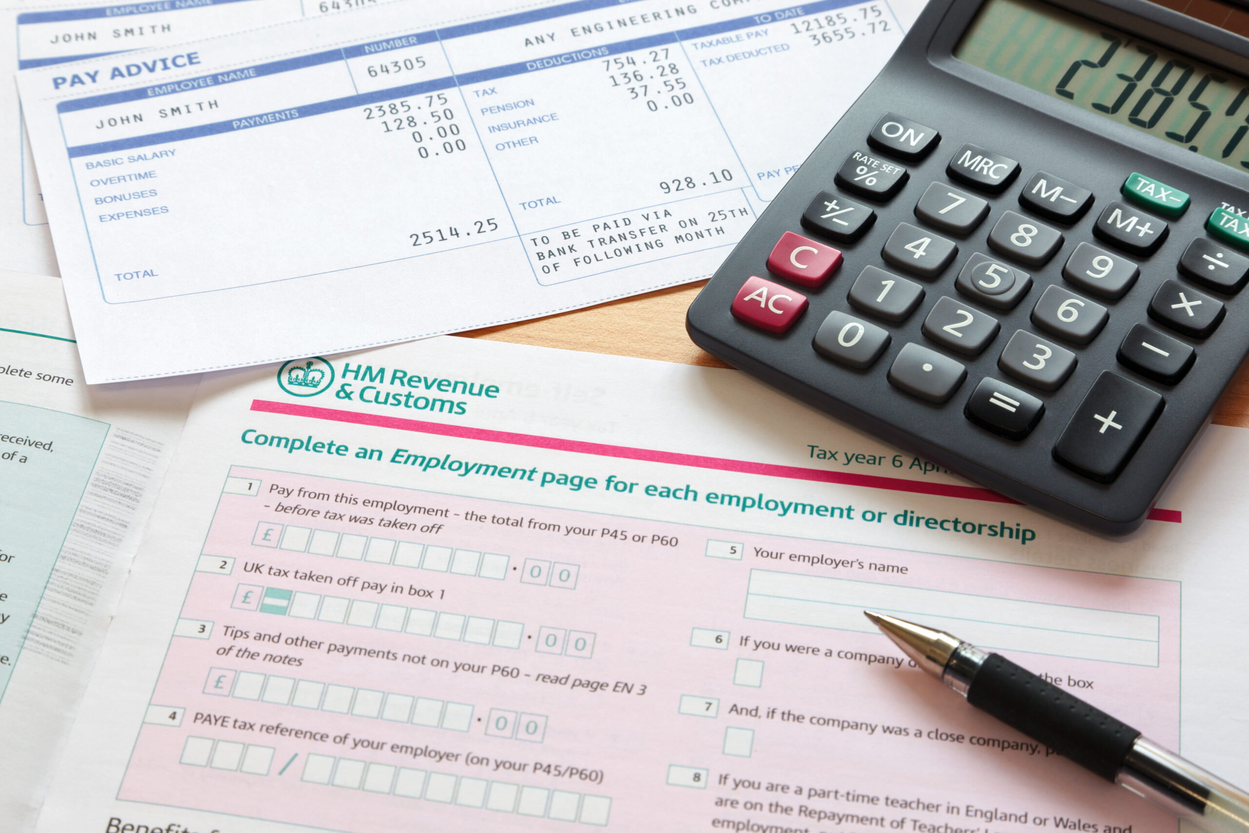 Photo of a UK self assessment tax return with calculator and payslips. The payslip is a mock up the names and all other information on it is fictional.
